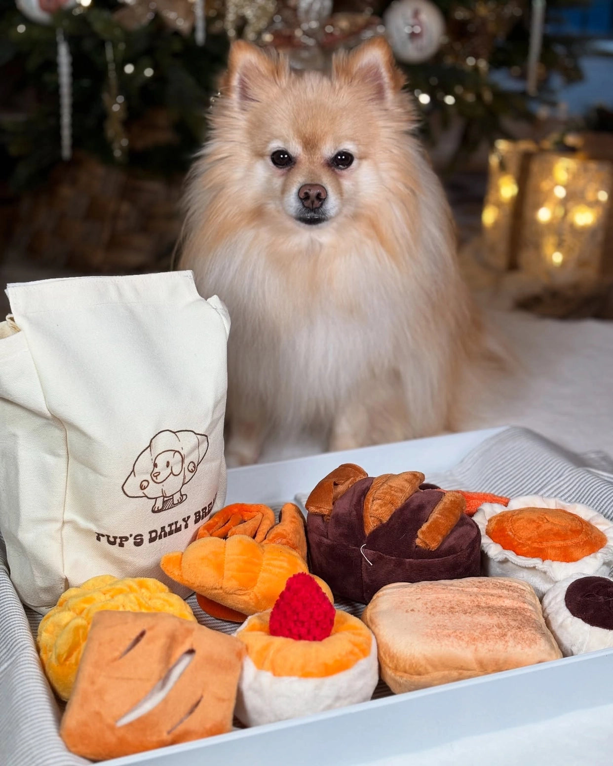 A fluffy tan Pomeranian sits behind a tray of Nocciola 10-Pack Crinkle & Squeaky Bread Dog Toys with Treat Bag, shaped like pastries and bread. A decorated Christmas tree with glowing lights twinkles in the background.