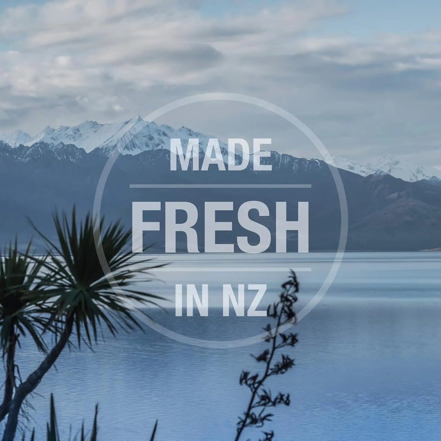 A tranquil lake with snowy mountains and native plants, overlaid with “MADE FRESH IN NZ” in bold white letters within a circle, showcases that Feline Natural Lamb Feast Grain-Free Canned Cat Food uses ethically sourced ingredients.