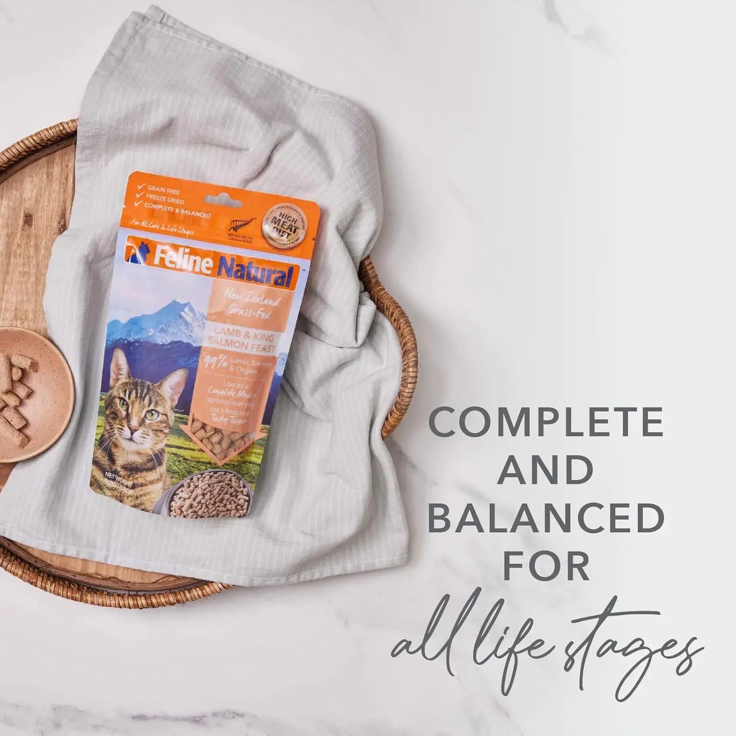 A bag of Feline Natural New Zealand Beef & Hoki Feast Grain-Free Freeze-Dried Cat Food sits on a beige cloth in a wicker tray beside a bowl, with text stating its complete and balanced for all life stages.