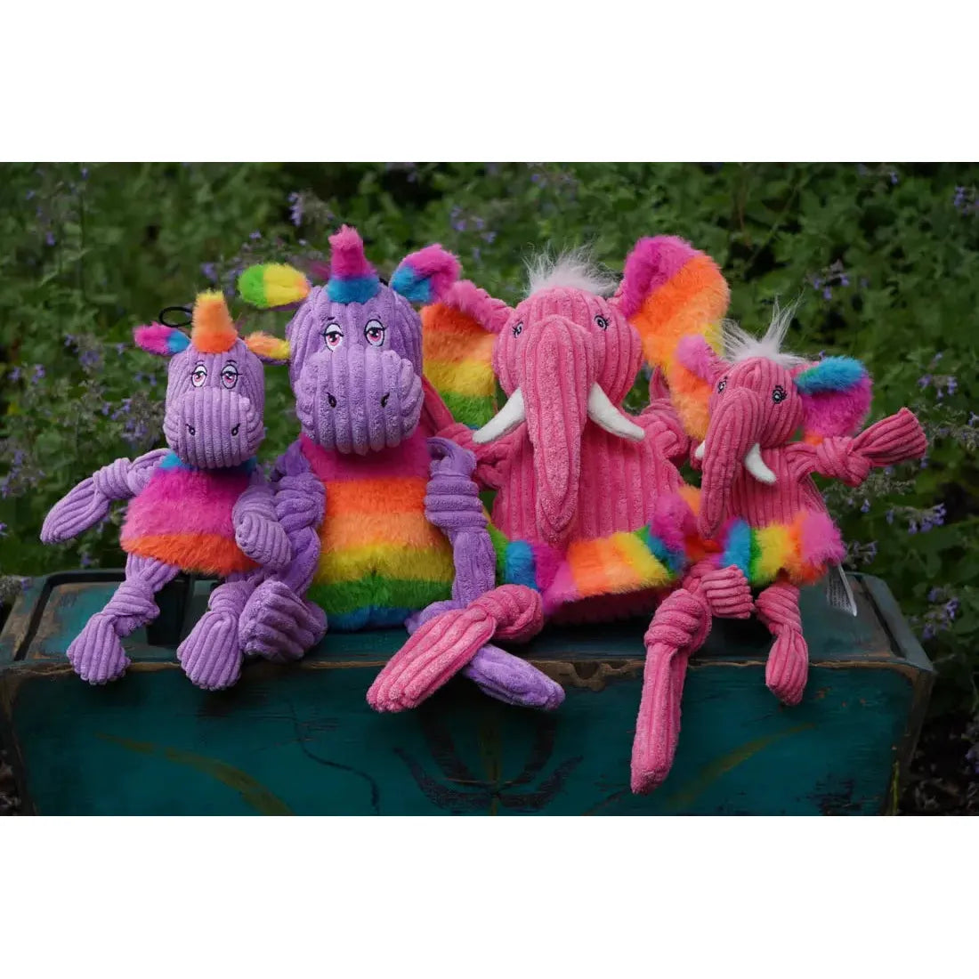 Four HuggleHounds Rainbow Durable Plush Corduroy Knotties Squeaky Dog Toys—two elephant and two unicorn designs—made with ribbed corduroy and bright rainbow stripes, sit on a teal bench with greenery in the background.