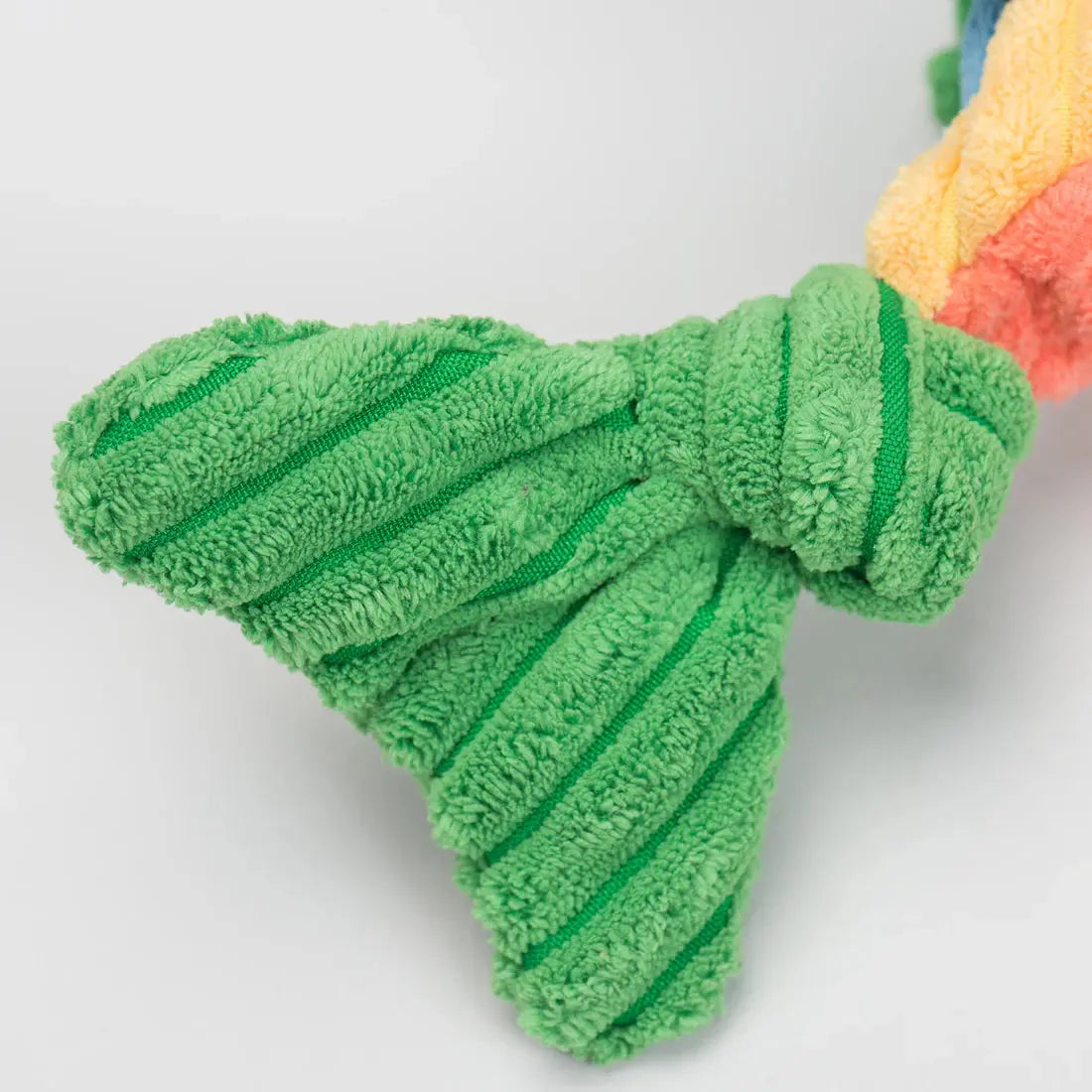 Close-up of a green, textured plush bird foot with visible stitching and ribbing, part of the HuggleHounds Raucous Rainbow Trout Knottie Tough Squeaky Plush Dog Toy. The multicolored plush toy’s edge appears in the background.