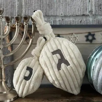 Two HuggleHounds Shining Collection Dreidel Durable Plush Dog Toys with Hebrew letters sit on a wooden surface near a silver menorah and ornament, with Star of David decorations and a brick wall in the background.