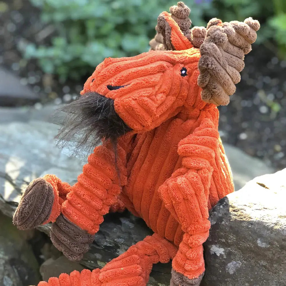 The HuggleHounds Woodlands Durable Plush Corduroy Knottie Moose Squeaky Dog Toy features textured orange fur and brown antlers, shown sitting outdoors on a rock amid greenery.