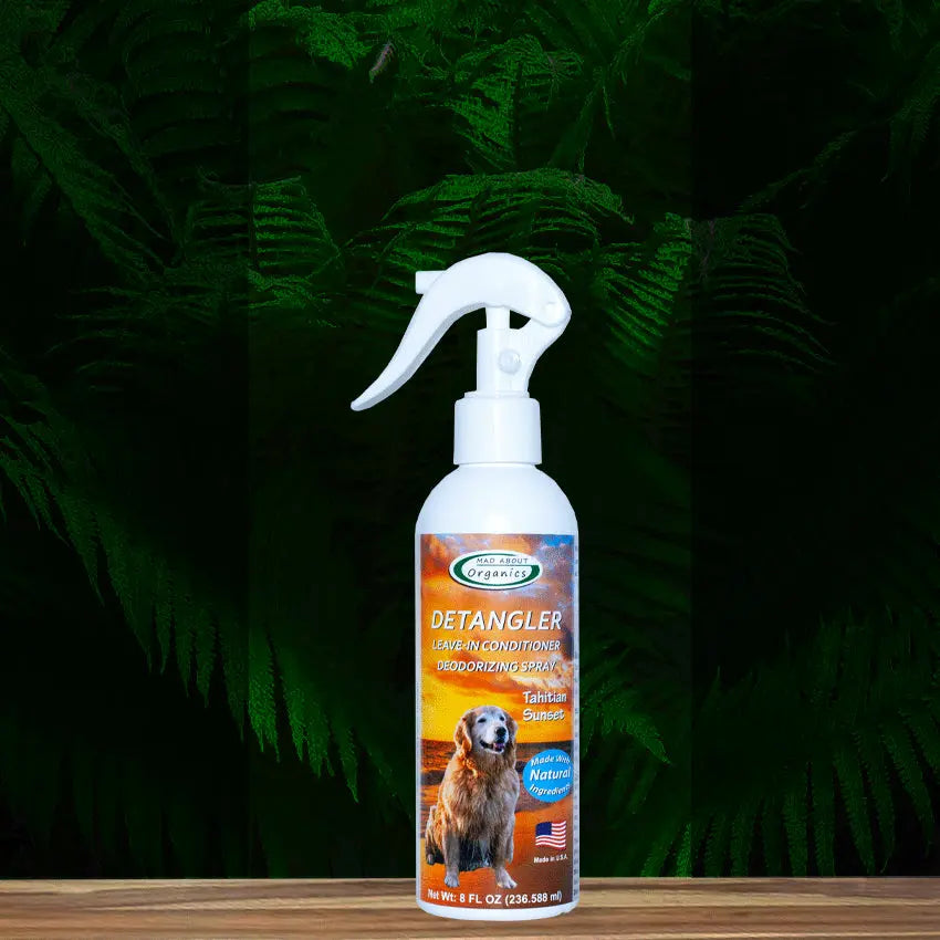 An 8oz bottle of Mad About Organics Detangler Spray for pets sits on a wooden surface with green fern leaves in the background. This natural dog conditioner features a fresh cedarwood scent and a label with a dog and American flag.