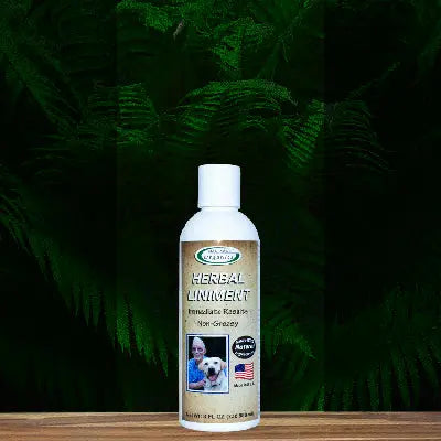 A bottle of Mad About Organics Herbal Relief Liniment with a white cap rests on wood among green ferns. The label features a person, a dog, and an American flag, highlighting natural ingredients like Arnica.