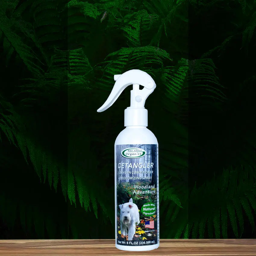 A white-nozzle bottle of Mad About Organics Detangler Spray - Knot-Free Coats Made Easy 8oz with a cedarwood scent sits on wood before green ferns. The label features a dog, “Woodland Adventure,” and “Made with Natural Ingredients.”.