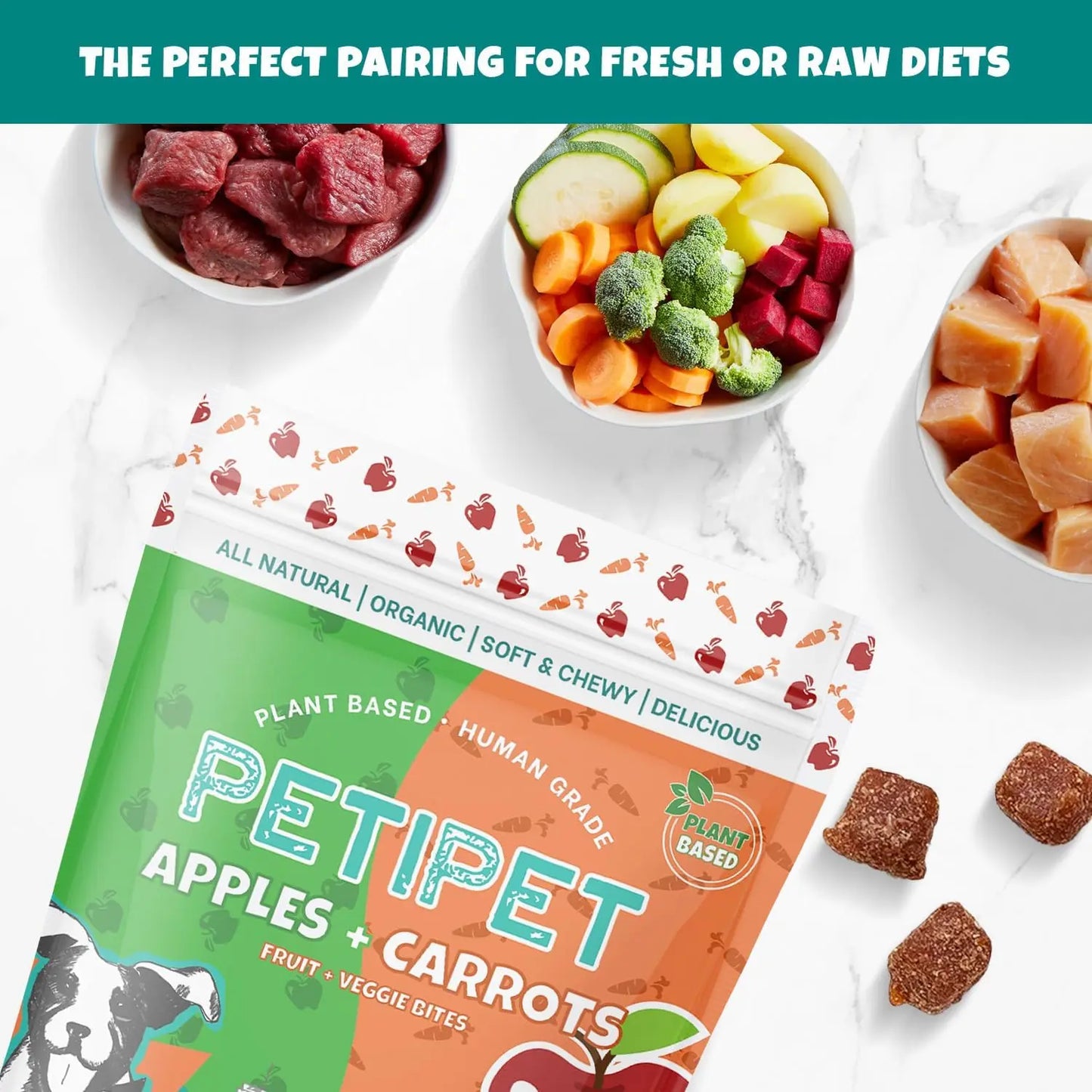 A 5 oz bag of PETIPET Apples + Carrots Bites Soft & Chewy Dog Treats is shown on a marble surface among bowls of veggies, fruit, and meat. These eco-friendly, plant-based treats are perfect for fresh or raw diets.