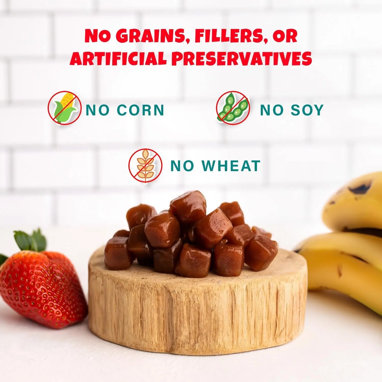 A pile of PETIPET Bananas + Strawberries Bites Soft & Chewy Dog Treats sits on a wooden platform with a strawberry and bananas. Text above reads: No grains, fillers, artificial preservatives, corn, soy, or wheat—with related icons.