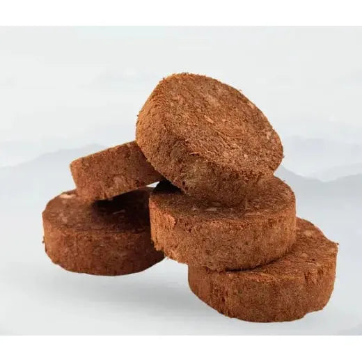 Four round, brown, coarse-textured discs of Rocky Mountain Raw Premium Freeze-Dried Raw Meal for Dogs Beef Formula 14 oz are stacked in a pyramid against a plain, light background.