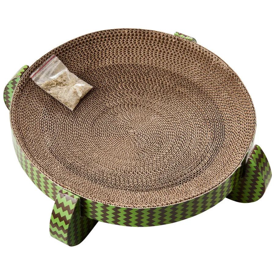 The Spot Nest Cat Scratcher by Spot is a round cardboard scratcher with green zigzag sides, designed to support nail health. It includes a catnip packet on the surface and stands on four short legs.