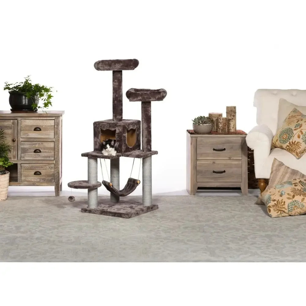 The Prevue Pet Products Kitty Power Paws Plush Party Tower by Prevue Pet, featuring scratching posts, platforms, a hammock, and a cubby, stands in a living room with gray furniture, green plants, and patterned pillows on a beige sofa.