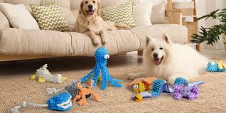 Two dogs relax in a living room, surrounded by various colorful stuffed animal toys scattered on a rug in front of a beige sofa with patterned cushions.