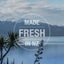 A tranquil lake with snowy mountains and native plants, overlaid with “MADE FRESH IN NZ” in bold white letters within a circle, showcases that Feline Natural Lamb Feast Grain-Free Canned Cat Food uses ethically sourced ingredients.