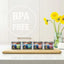 Four cans of Feline Natural Lamb Feast Grain-Free Canned Cat Food by Feline Natural are arranged on a wooden board in a kitchen, with a BPA FREE label and a vase of yellow flowers in the background, emphasizing quality and care.
