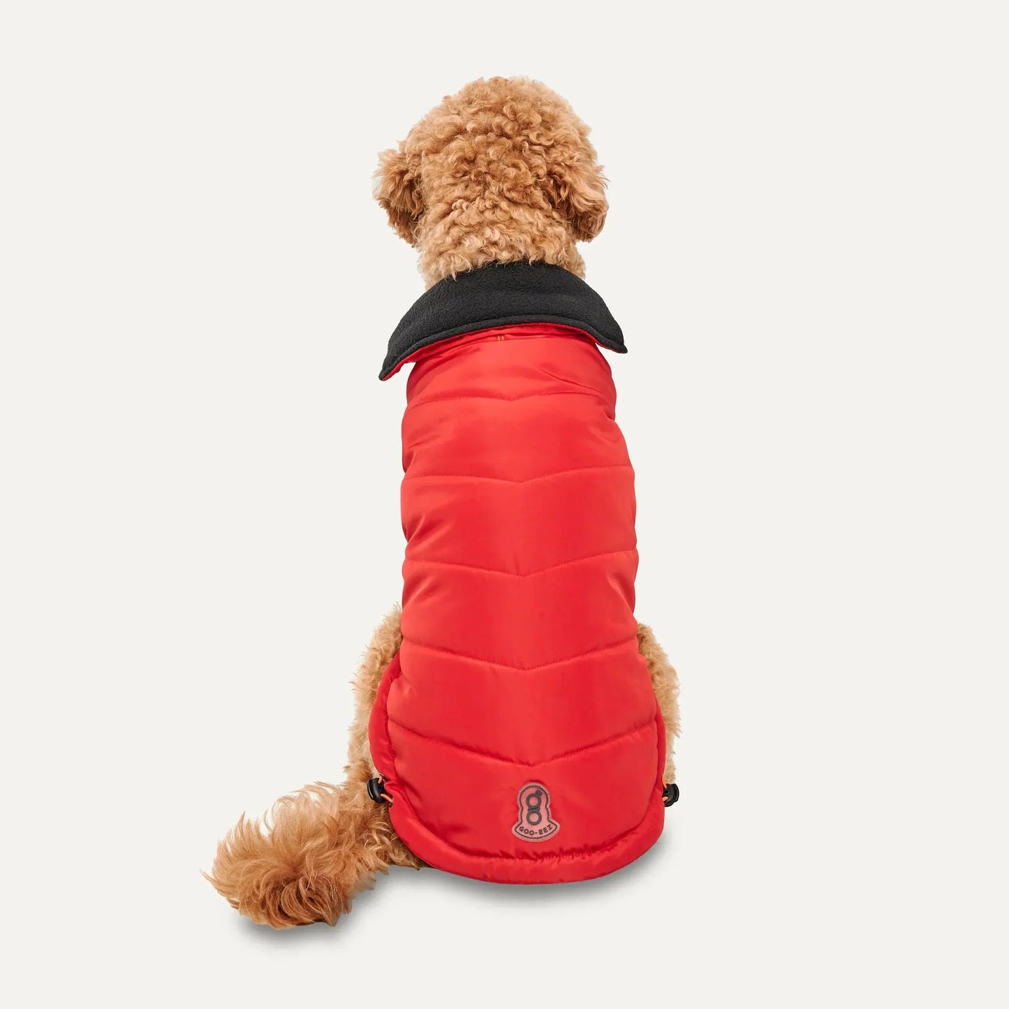 A small, curly-haired dog faces away, wearing the Goo-Eez Mock Neck Signature Dog Jacket by Goo-Eez. The bright red jacket has a black collar, a patch, and two adjustable cords at the bottom hem. Background is plain white.