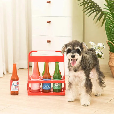 A happy dog stands beside a red carrier holding the Hugsmart Pet Boozy Tailz Heinecanine TPR Rubber Dog Toy in a bright room with light wood floors, a white cabinet, and a potted plant in the background.