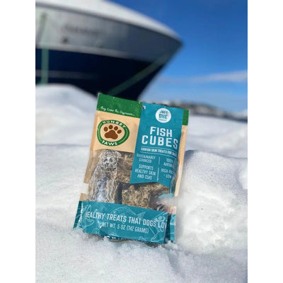 A 5 oz bag of Hungry Paws Premium Cod Skin Cubes Dog Chew Treats stands upright on snow, with a boat and blue sky behind. The packaging highlights sustainability, natural ingredients, and dental health support for dogs.