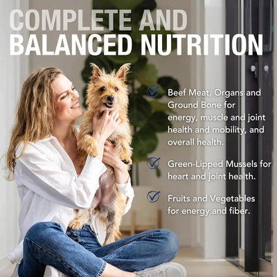 A smiling woman in a white shirt holds a happy small dog indoors. Text highlights K9 Natural Grass-Fed Beef Feast Grain-Free Canned Dog Food with wholefood ingredients: grass-fed beef for energy, mussels for joint health, and fruits and veggies for fiber.