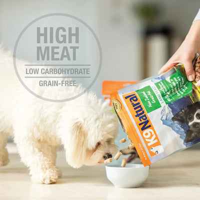 A small white dog eats kibble from a bowl while a person pours more food from a K9 Natural Hoki & Beef Grain-Free Freeze-Dried Dog Food bag. Text on the image reads: High meat, low carbohydrate, grain-free.