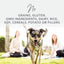 A happy dog runs on grass toward the camera, two women in the background. Text above features K9 Natural Hoki & Beef Grain-Free Freeze-Dried Dog Food Topper: no grains, gluten, GMOs, dairy, rice, soy, cereals, potato or fillers.