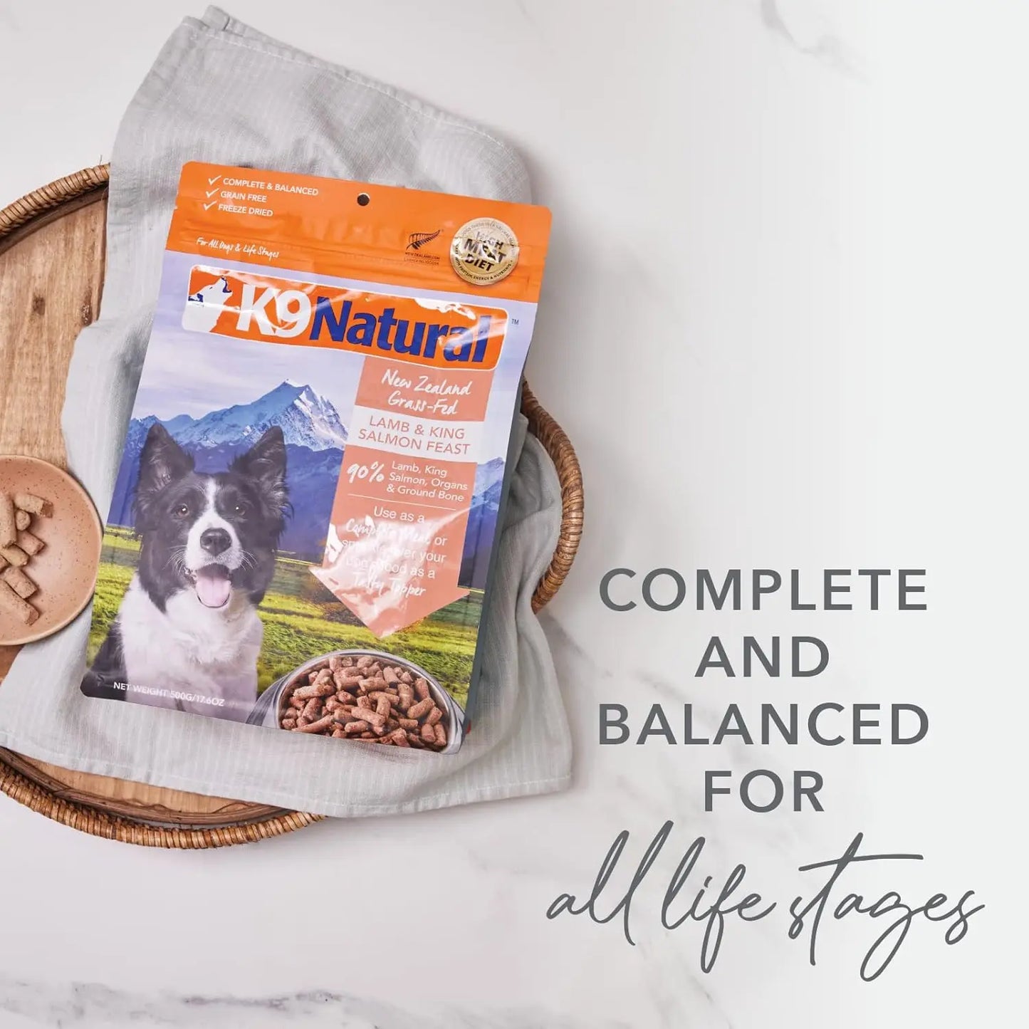 A package of K9 Natural Hoki & Beef Grain-Free Freeze-Dried Dog Food Topper rests on a gray cloth in a basket. The text reads: Complete and balanced for all life stages. Dog treats and a paw-shaped biscuit are nearby.