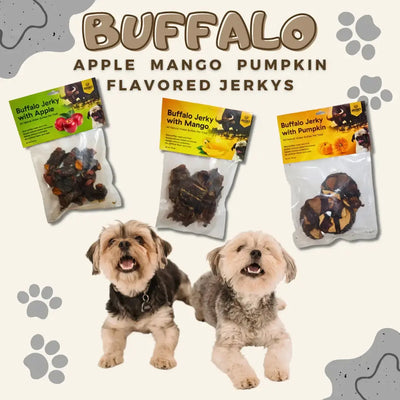 Two small fluffy dogs sit before three hanging Advance Pet Products Long Lasting Water Buffalo Jerky Treats in apple, mango, and pumpkin flavors. Paw prints and text emphasize the all natural ingredients for healthy dog chewing.