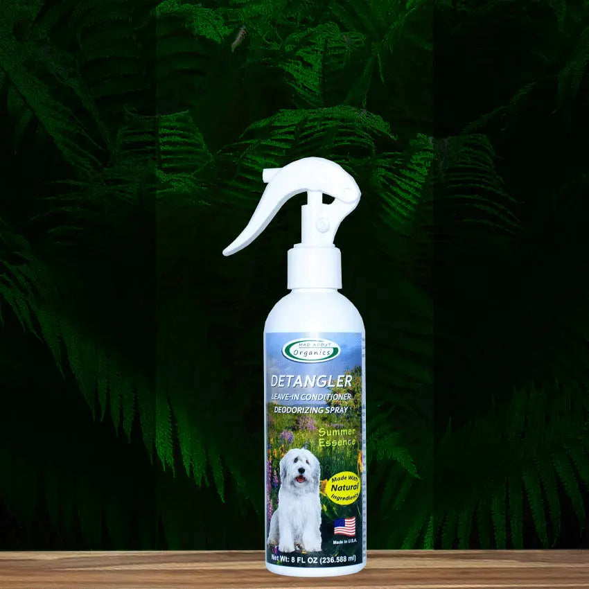 An 8oz bottle of Mad About Organics Detangler Spray—Knot-Free Coats Made Easy—resting on a wooden surface with green ferns. The label features a white dog and promotes its deodorizing detangler spray formula.