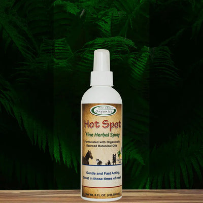 An 8oz white spray bottle of Mad About Organics Herbal Hot Spot Spray – Natural Relief for Pets sits on a wooden surface with green ferns behind it. The label features organic ingredients and shows silhouettes of cats and dogs.