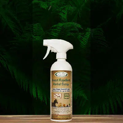 A spray bottle of Mad About Organics Herbal Spray – Natural Bug Repellent for Pets sits on a wooden surface among green ferns, with a label featuring a horse and dog and its natural insect repellent benefits.