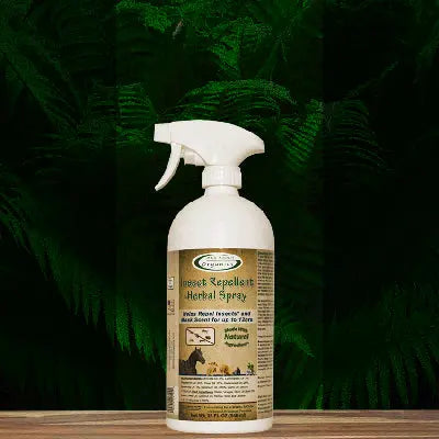 A white spray bottle of Mad About Organics Herbal Spray – Natural Bug Repellent for Pets rests on a wooden surface with leafy ferns behind it. Its beige label displays animal silhouettes and green text highlighting safe, natural ingredients.