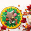 The Nocciola Colorful Turkey-Themed Dog Toys sit on a round yellow and green mat with plush veggies and leaves, surrounded by autumn leaves, berries, and a white pumpkin—ideal for puppies and small breeds.
