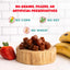 A pile of PETIPET Bananas + Strawberries Bites Soft & Chewy Dog Treats sits on a wooden platform with a strawberry and bananas. Text above reads: No grains, fillers, artificial preservatives, corn, soy, or wheat—with related icons.
