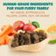 A wooden spoon with brown pet treats sits among carrots, apples, and orange slices. Text reads: PETIPET Bladder Booster Cranberry Chews for Dogs—urinary and kidney health support. No GMOs, byproducts, fillers, corn, soy, or wheat.