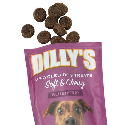A purple bag of Poochie Butter Dillys Blueberry Soft & Chewy Dog Treats is open, with several round, brown treats spilling out. The bottom of the bag shows part of a dogs face.