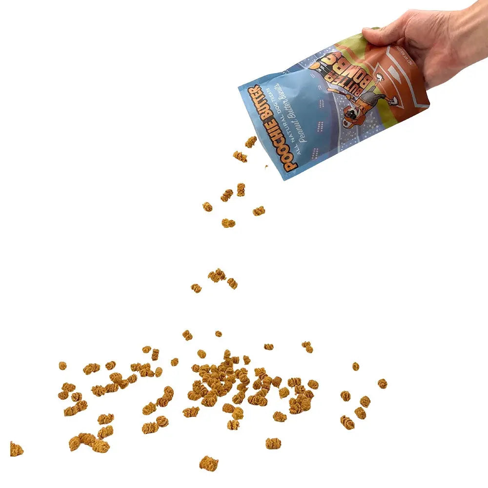 A hand pours Poochie Butter Peanut Butter Crunchy Dog Treats, 8oz, from a blue Poochie Butter bag onto a white surface, scattering the small brown treats.