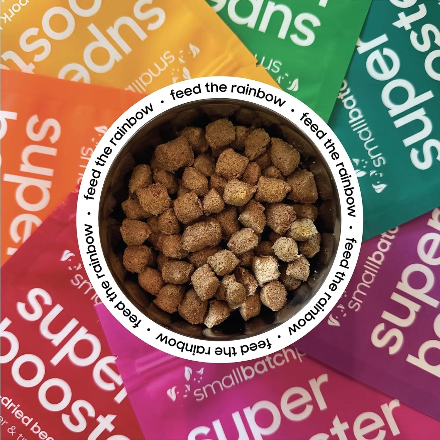 A bowl of pet food is surrounded by colorful packets of Smallbatch Super Booster Chicken SmallBites Freeze-Dried Dog & Cat Meal Topper & Treats 18oz, with the bowl’s rim repeating “feed the rainbow” to highlight their single-source protein goodness.