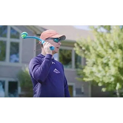A person in sunglasses, a red cap, and a dark long-sleeve shirt uses the Spunky Pup Squeak & Throw Ball Launcher for Dogs to pick up a tennis ball hands-free outdoors near a house and trees. Brand: Spunky Pup.