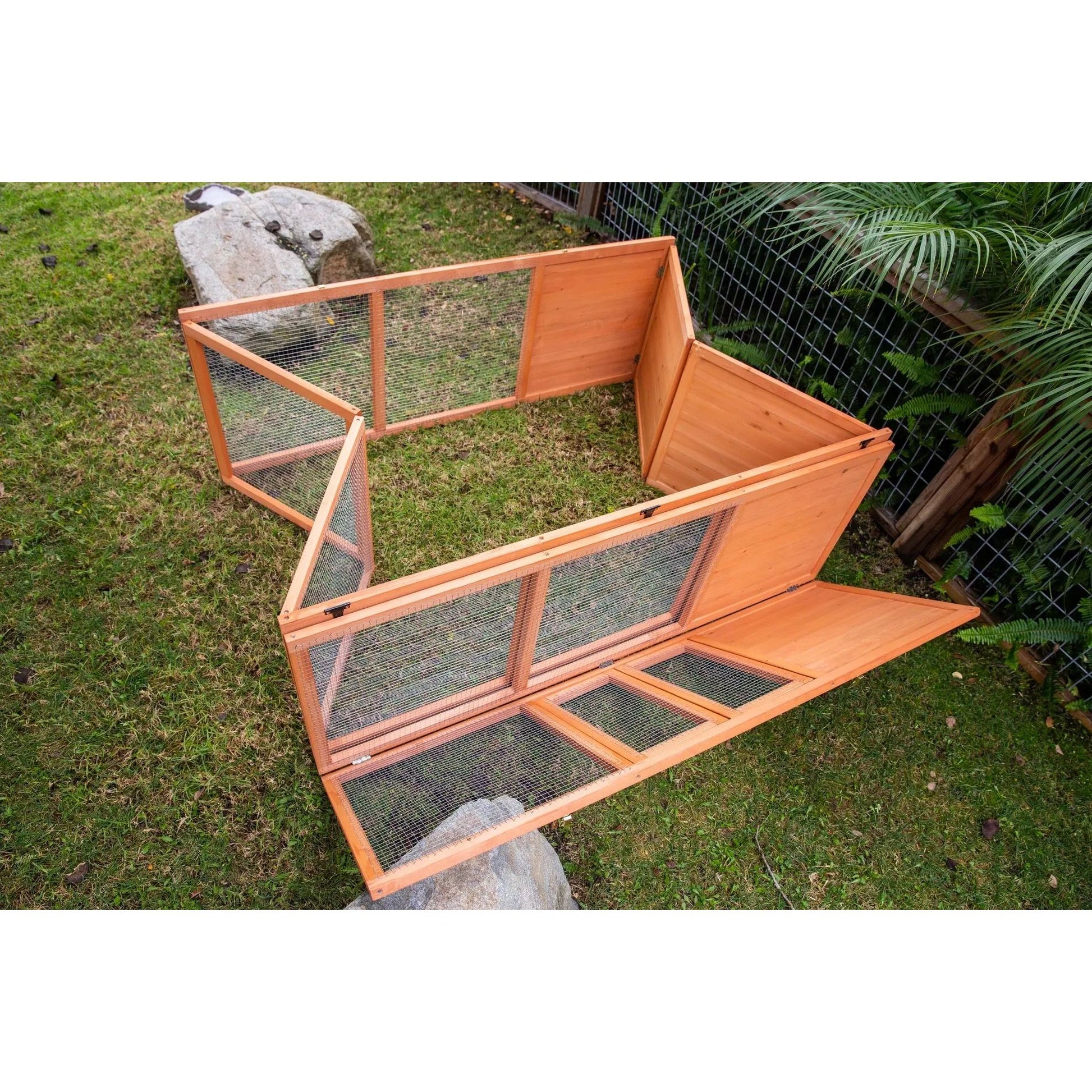 A partially assembled Zoo Med Laboratories Easy-Fold Tortoise Terrarium Yard sits on backyard grass, featuring open wooden and wire panels and doors, with nearby rocks and plants visible.