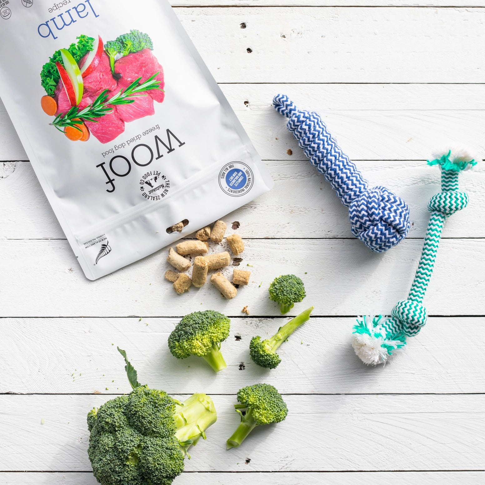 Woof lamb freeze dried dog food bag with broccoli and dog toys on a white wooden surface